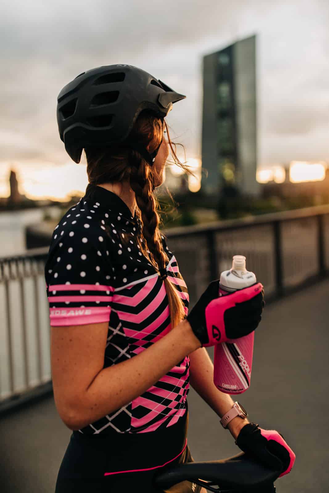 Frau steht mit Trinkflasche und Fahrradbekleidung auf Brücke und schaut auf den Sonnenuntergang